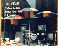 Photographie couleur d'une vitrine de magasin dans laquelle sont exposées des télévisions sous des parasols. Sur la vitrine, on lit "Au premier étage, salon haute fidélité, meubles radio-phono, TV couleur, orgues électroniques".