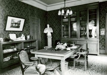 Photographie noir et blanc du bureau du recteur de l'université de Lille. Un téléphone et divers papiers sont posés sur une grande table au milieu de la pièce. De chaque côté sont disposées des bibliothèques ; on remarque un buste au fond de la pièce. 