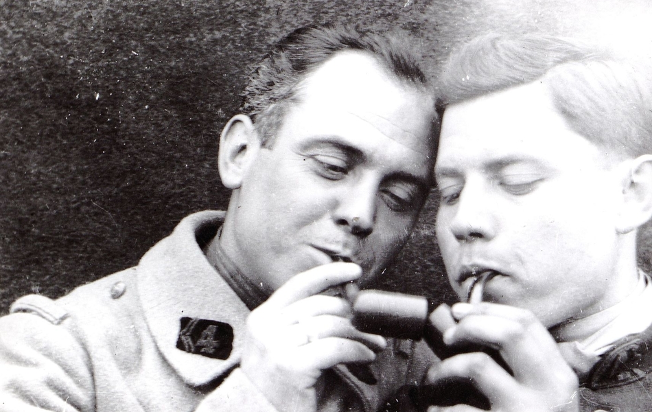 Photographie noir et blanc montrant deux hommes tête contre tête, l'un en train d'allumer sa pipe sur celle de son voisin.
