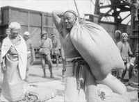 Photographie noir et blanc montrant au premier plan un asiatique, torse nu, portant un sac sur son épaule. À l'arrière plan, d'autres hommes devant un wagon de marchandises.