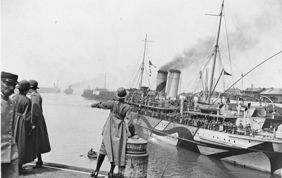 Photographie noire et blanc montrant une femme de dos, sur un quai, devant un navire amarré, peint de zébrures.
