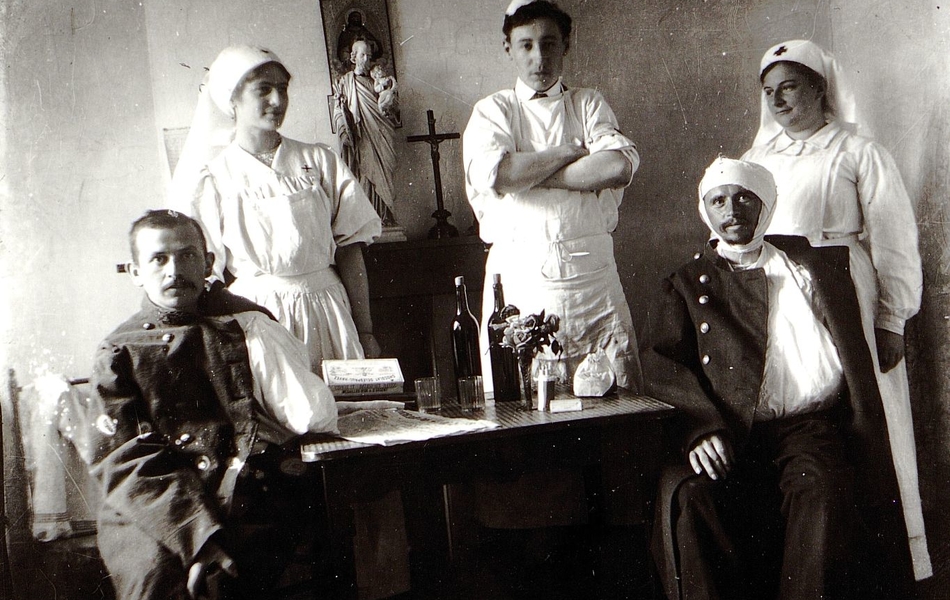 Photographie noir et blanc montrant deux soldats blessés et couverts de bandage assis de part et d'autre d'une table sur laquelle sont posées une bouteille de vin et une boîte de chocolat. Autour d'eux, un chirurgien avec un tablier et deux infirmières derrière un crucifix fixé au mur.