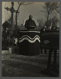 Photographie noir et blanc montrant un homme faisant un discours sur une tribune. Au premier plan un cerceuil.