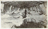 Photographie noir et blanc montrant l'intérieur d'un hôpital monté dans un baraquement en bois. Une double rangée de lits se fait face ; au milieu, debout, se tient un homme.