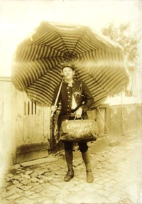 Photographie sepia montrant un soldat, une pipe dans la bouche, tenant une valise et un parasol ouvert dans l'autre main.