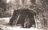 Photographie noir et blanc montrant un abri en bois recouvert de végétation. À l'intérieur, un homme et des obus.