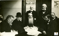 Photographie noir et blanc montrant sept hommes assis autour d'une table recouverte de papiers.