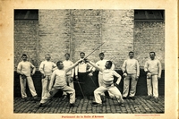 Photographie noir et blanc montrant deux épéistes en plein duel. Derrière eux posent sept hommes devant un mur de briques.