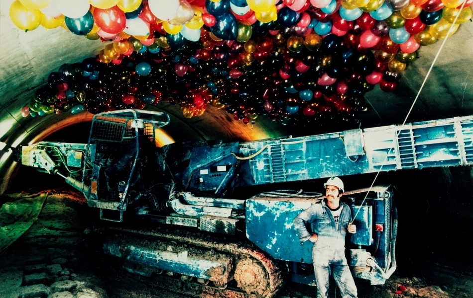 Photographie couleur montrant un homme en tenue d'ouvrier posant devant une machine de chantier, le regard tourné vers des centaines de ballons de baudruche colorés ammassés au-dessus de lui dans un tunnel.