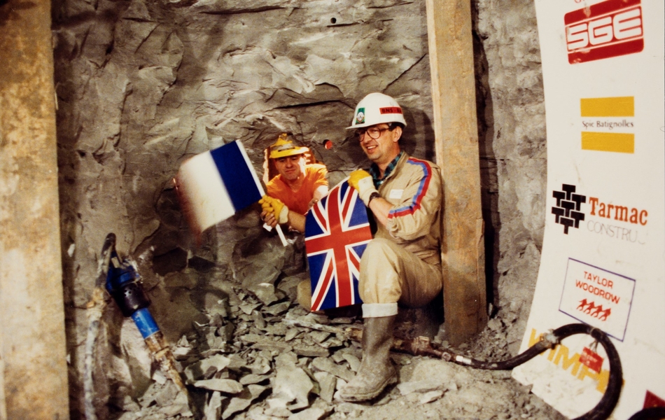 Photographie couleur montrant deux hommes de par et d'autre un tunnel percé, s'échangeant des drapeaux français et britanniques.