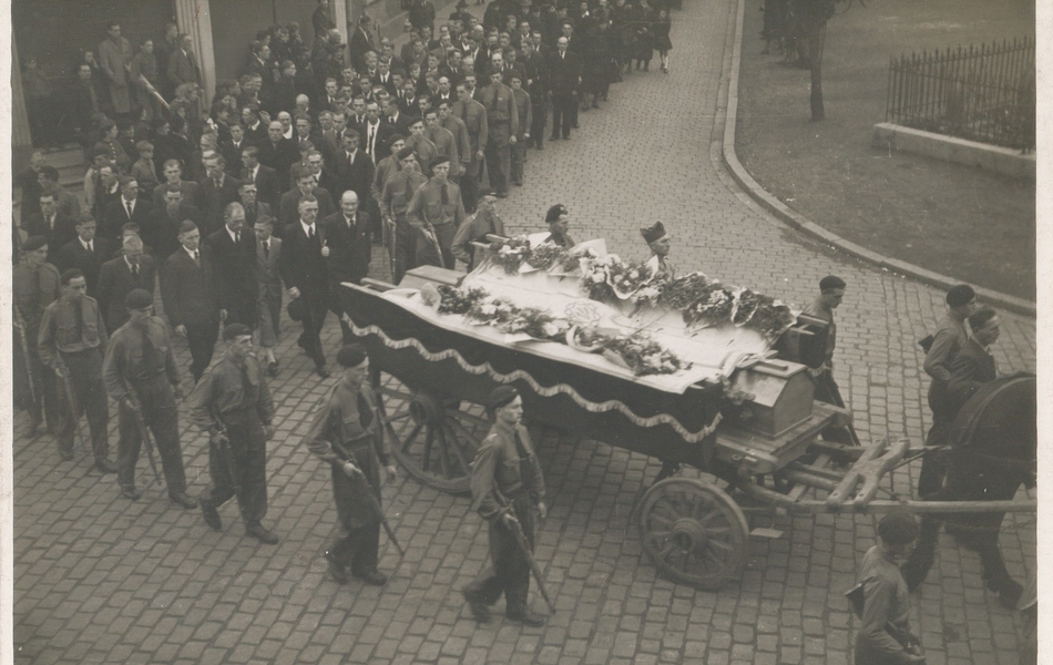 Photographie noir et blanc montrant des hommes portant un cercueil dans une rue, suivis d'une procession.