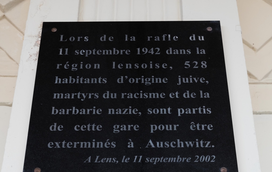 Photographie couleur d'une plaque commémorative sur laquelle on lit : "Lors de la rafle du 11 septembre 1942 dans la région lensoise, 528 habitants d'origine juive, martyrs du racisme et de la barbarie nazie sont partis de cette gare pour être exterminés à Auschwitz. A Lens, le 11 septembre 2002".