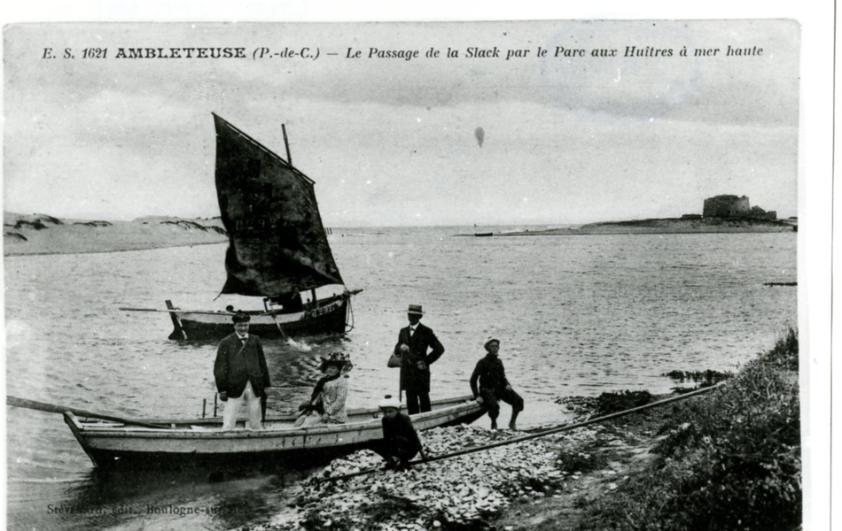 Carte postale en noir et blanc d’un bras de mer dans l’estuaire de la Slack. Au premier plan, une barque accostée à un rocher, avec à son bord une femme assise et deux hommes debout. À côté de cette barque, deux enfants portent un béret à pompon. Derrière, un cat-boat animé traverse la carte postale