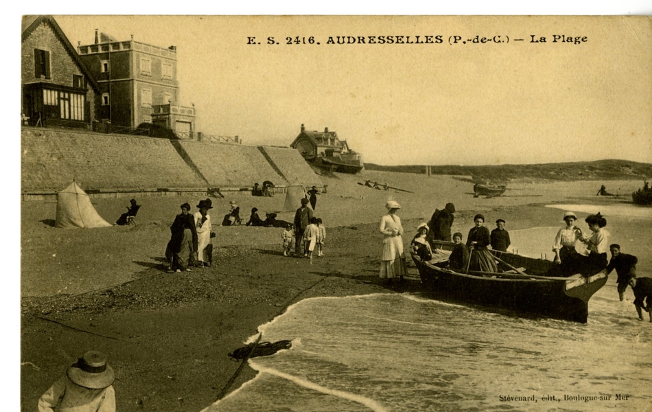 Carte postale en noir et blanc de la plage occupée par une vingtaine de personnes, dont une dizaine se trouve dans ou autour d’une barque