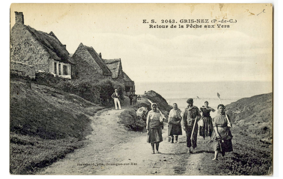 Carte postale en noir et blanc d’un chemin en terre battue menant à la plage. À gauche, se trouvent de petites maisons en hauteur. Un groupe de cinq pêcheurs munis de grands bâtons et de seaux remonte de la plage