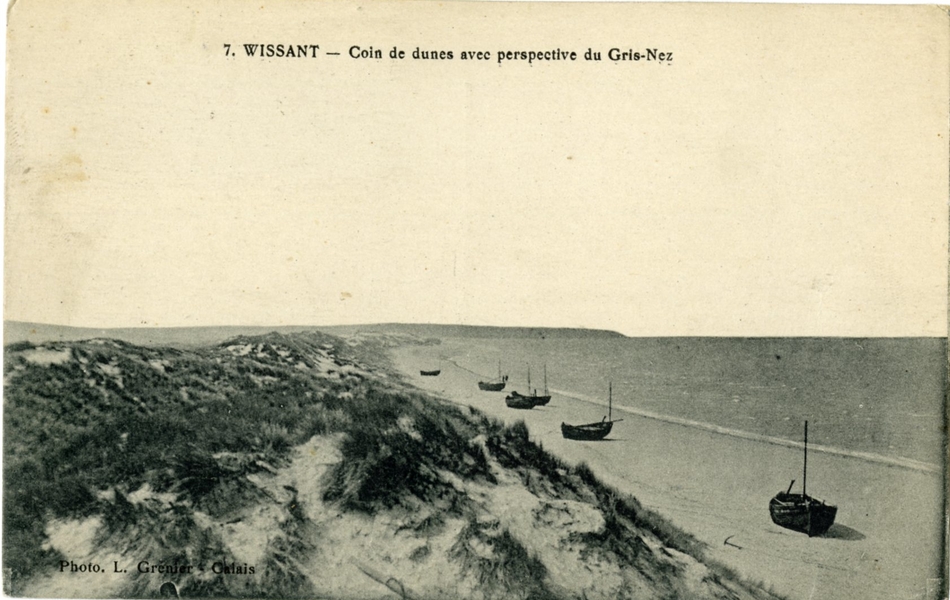 Carte postale en noir et blanc des dunes et de la plage, sur laquelle on aperçoit quelques barques échouées. En arrière-plan se dessinent les falaises du cap Gris-Nez