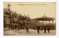 Carte postale noir et blanc d'une place où se dresse un kiosque à musique devant la façade de maisons. Au premier plan posent un jeune garçon et trois messieurs.