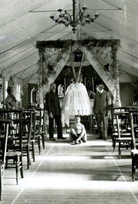 Photographie noir et blanc d'une cloche recouverte d'un drap brodé, encadrée par deux hommes de chaque côté et un garçon assis devant. Elle est exposée devant un autel, au bout d'une allée formée par des rangées de chaises.