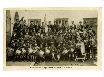 Photographie noir et blanc de l'ensemble de la fanfare composée d'une trentaine d'enfants de tous âges, vêtus de leur uniforme et tenant leurs instruments. Au centre, trois hommes et derrière eux, la bannière.