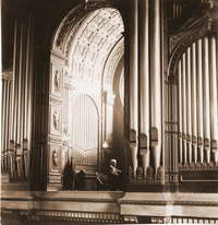 Photographie noir et blanc où on aperçoit un homme assis devant un clavier, entouré de grands tuyaux d'orgue.