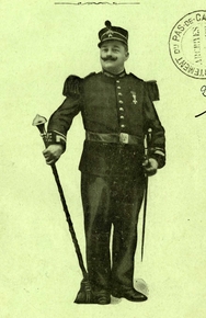 Photgraphie noir et blanc d'un homme posant en uniforme d'harmonie, une épée au côté. Il porte une médaille, un chapeau avec un petit panache, une grande moustache relevée et tient une pique dans sa main droite.