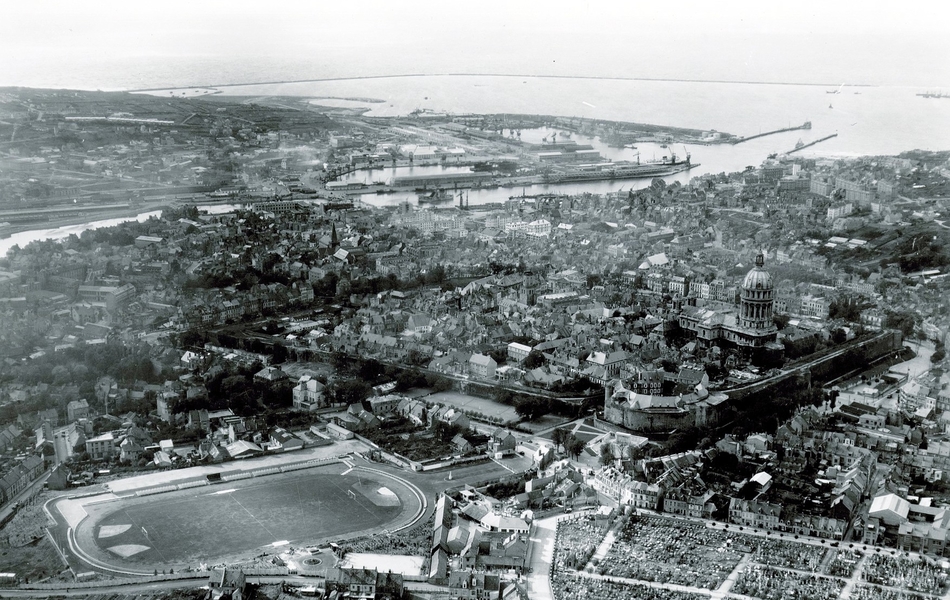 Photographie aérienne noir et blanc d'une ville où on voit un stade au premier plan.