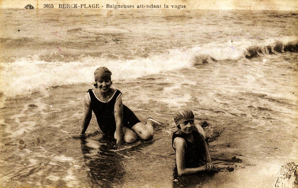 Carte postale noir et blanc de deux jeunes femmes en costume de bain portant un bonnet, assises sur le sable en bord de mer. Elles sourient au photographe