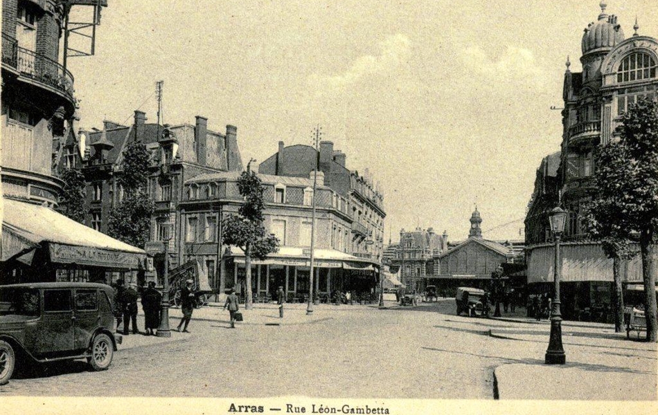 Carte postale noir et blanc représentant un croisement de la rue Gambetta au bout de laquelle on aperçoit l'entrée de la gare